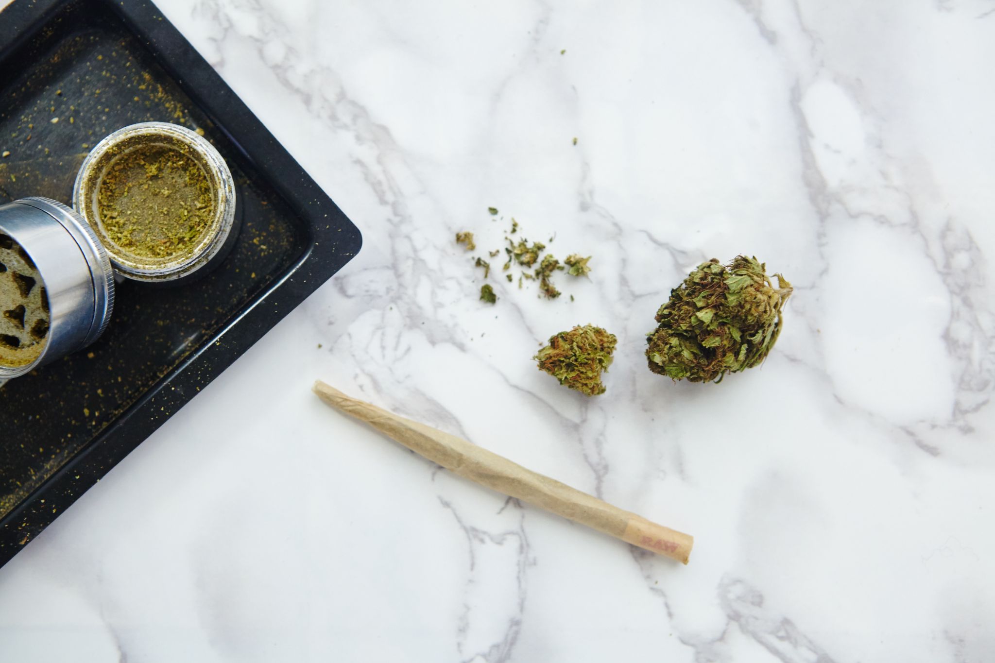 A flat lay photo of a black tray containing a metal grinder with ground cannabis, a small bud of cannabis, and a rolled joint on a marble surface.