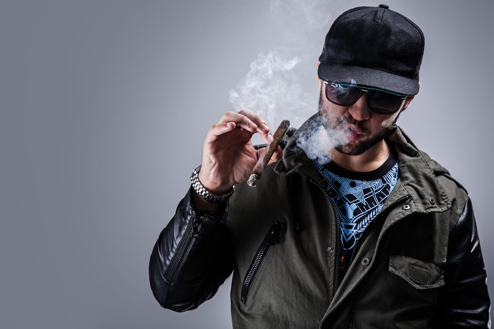 A man in a black leather jacket and a black baseball cap smokes a cigar and exhales smoke.