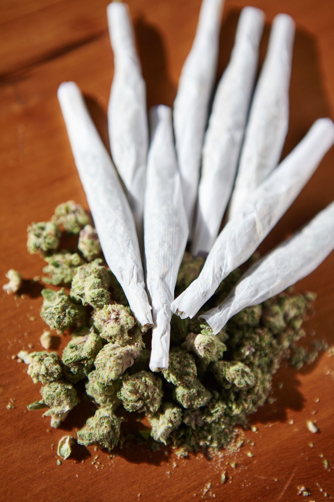  A pile of marijuana buds surrounded by pre-rolled joints on a wooden surface.